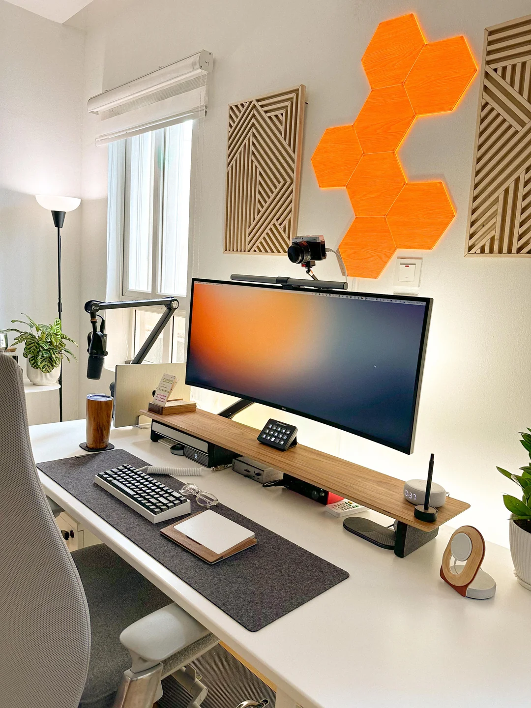 A modern, neatly organized desk setup with a curved monitor, keyboard, microphone, webcam, and ambient orange hexagonal wall lights. Plants and wooden decor add a stylish touch to the bright workspace.