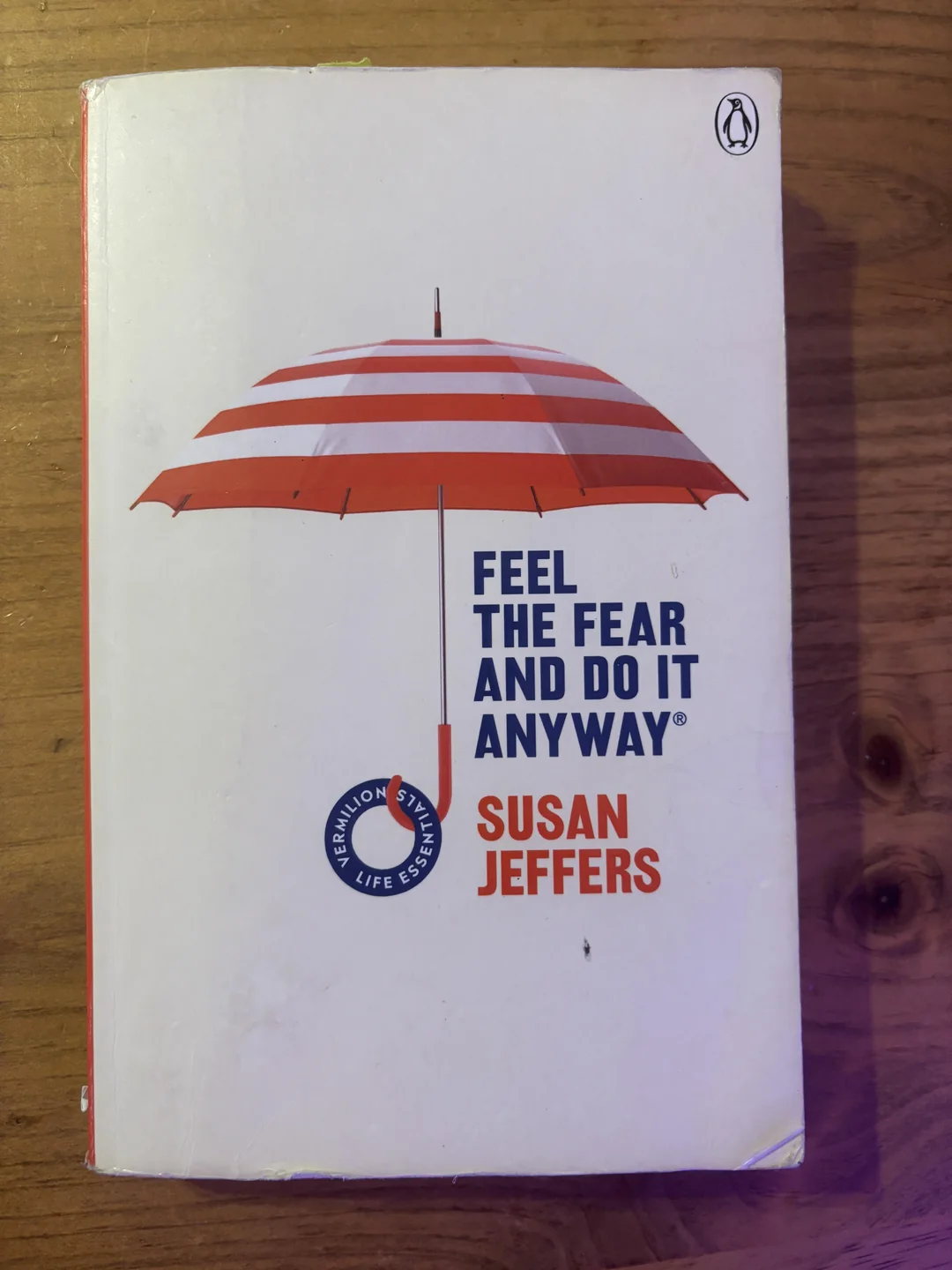 A book titled "Feel the Fear and Do It Anyway" by Susan Jeffers, featuring a red and white striped umbrella on a white cover, placed on a wooden surface.