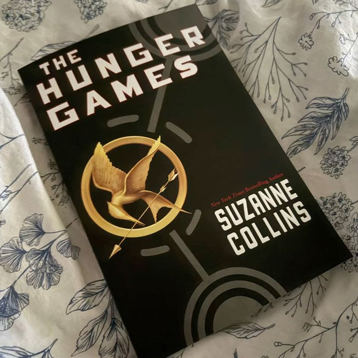 A copy of "The Hunger Games" by Suzanne Collins rests on a white fabric with a blue floral pattern. The book cover is black with a gold mockingjay pin and bold white lettering.