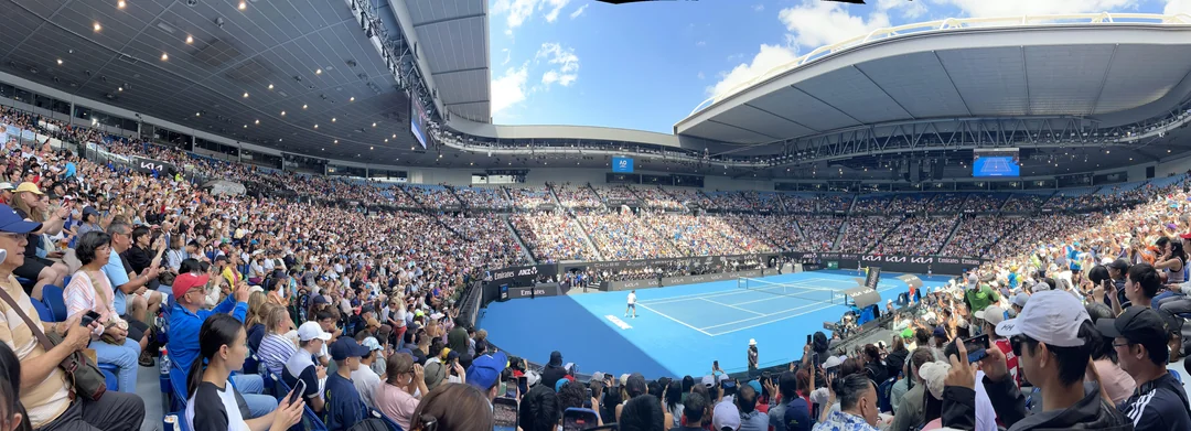 A packed stadium with thousands of spectators watching a tennis match on a blue court under a partly covered roof, with bright daylight and a clear sky visible above.