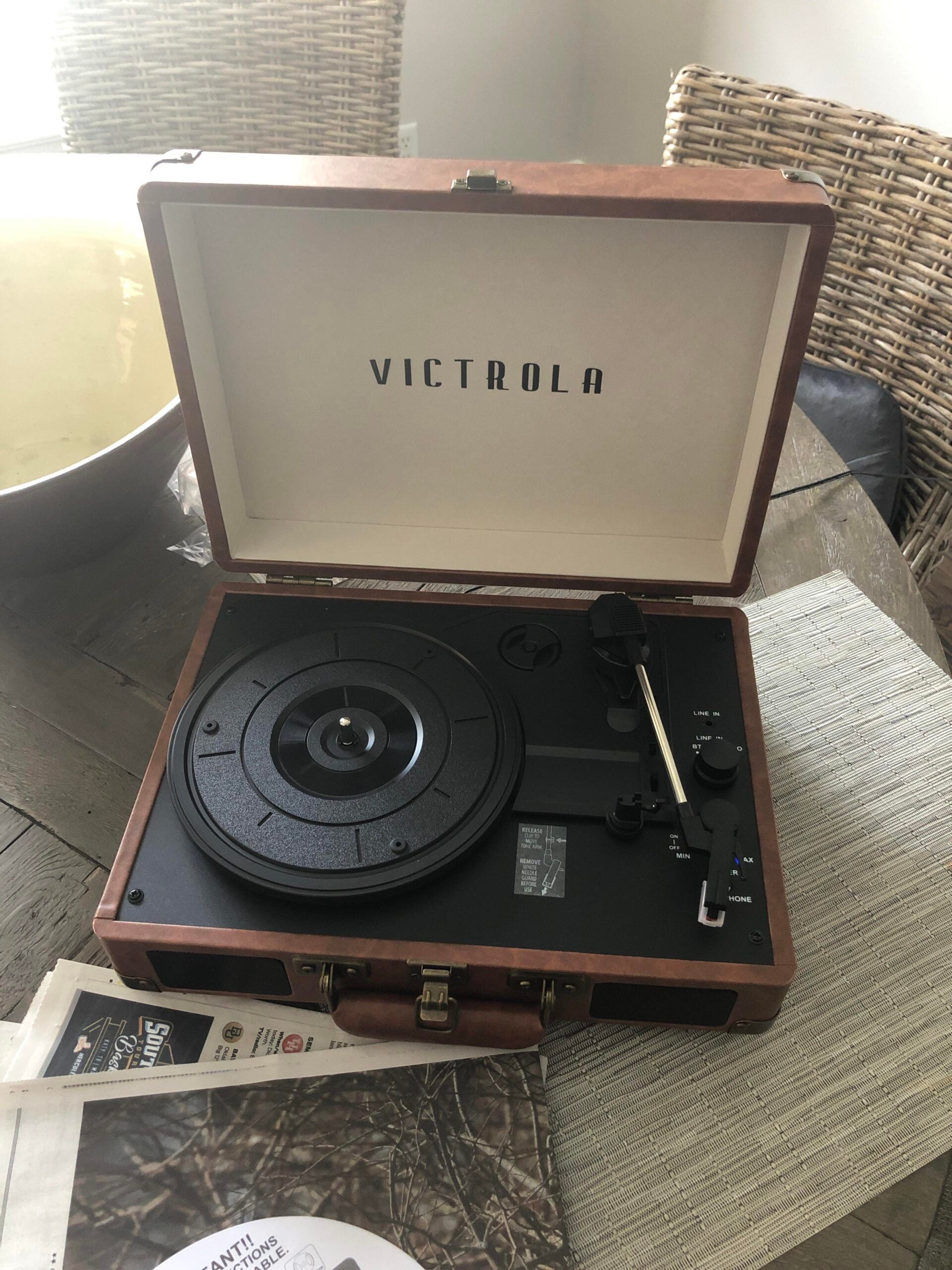 A brown Victrola portable record player sits open on a dining table with a wicker chair in the background and newspapers nearby. The tonearm rests on the player, ready for use.