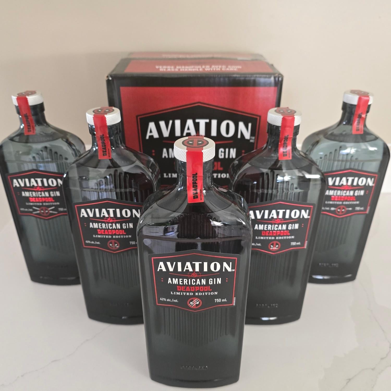 Five bottles of Aviation American Gin are arranged in front of a matching box, with all labels facing forward. The bottles have red caps and black labels with white and red text. The background is a plain light surface.