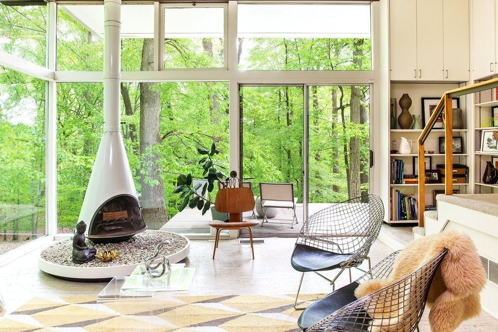 A bright, modern living room with floor-to-ceiling windows overlooking lush green trees. Features a white cone fireplace, contemporary chairs, a geometric rug, and a cozy patio outside. Shelving and stairs are visible on the right.