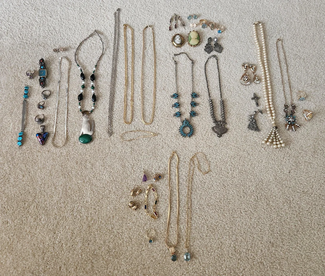 Assorted jewelry including necklaces, bracelets, rings, and earrings with various designs and colors, laid out on a beige carpet. Items include pearl strands, turquoise pieces, and silver and gold-toned accessories.