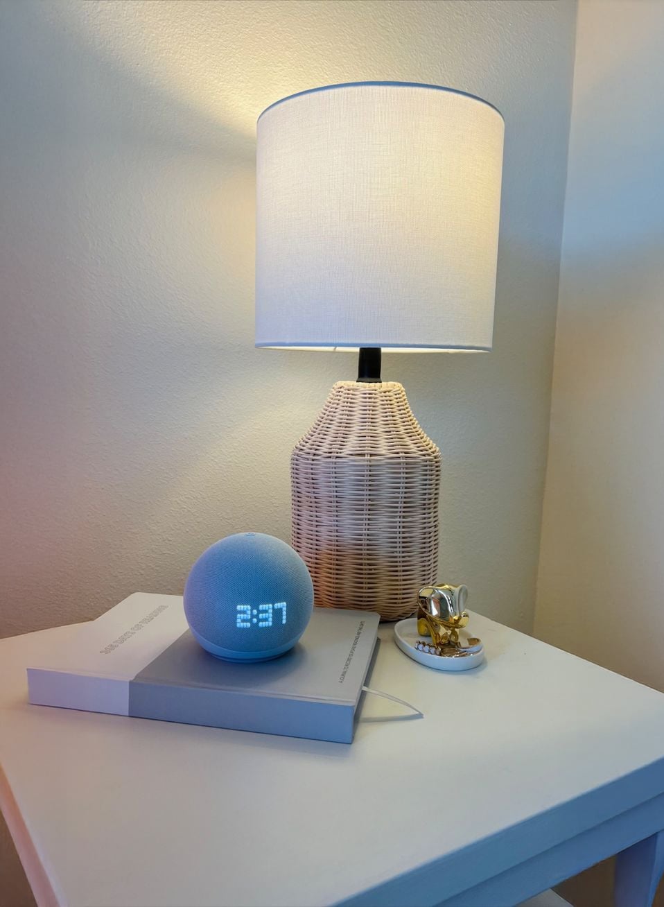 A bedside table with a woven lamp, a smart speaker displaying 3:37, two stacked books, and a small tray holding gold jewelry shaped like an animal.