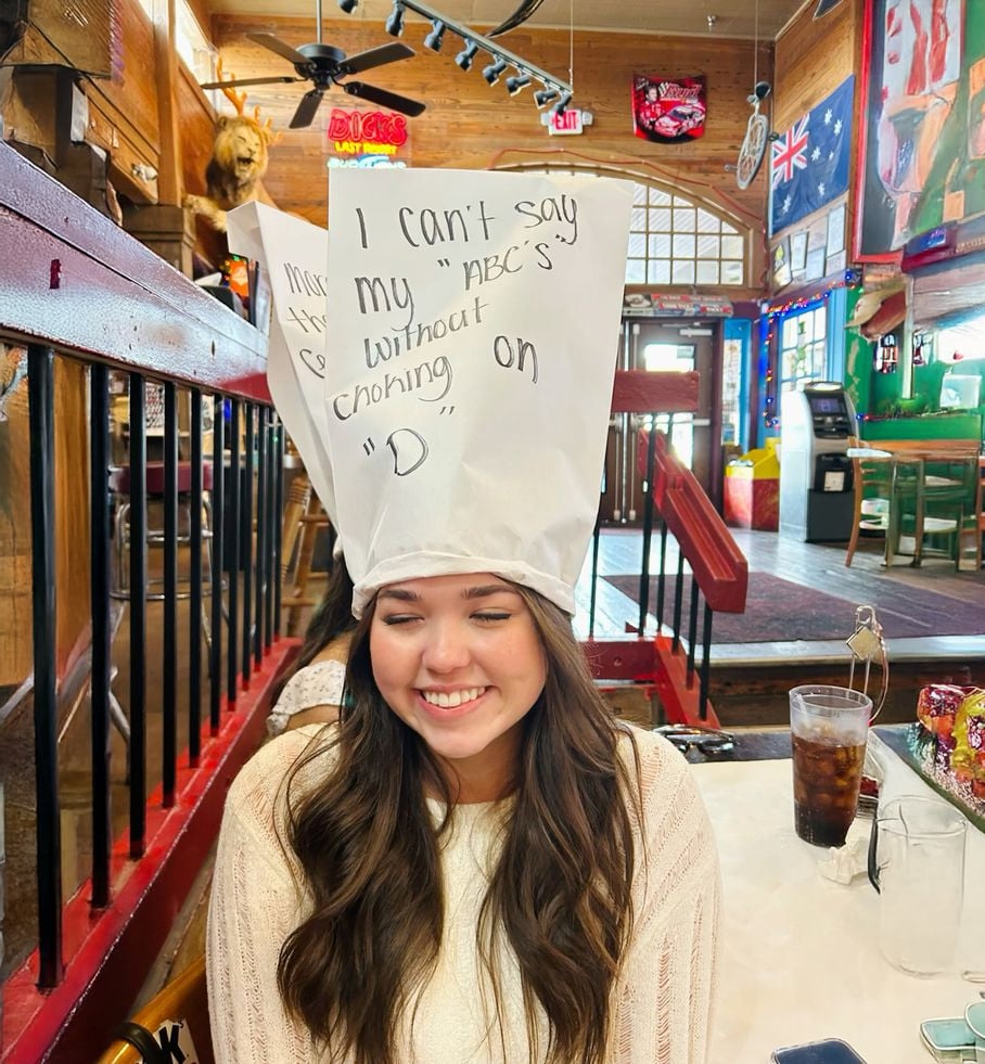 A young woman with long brown hair sits smiling at a restaurant table, wearing a large white paper hat with handwritten text that says, "I can't say my ABC's without choking on 'D'.