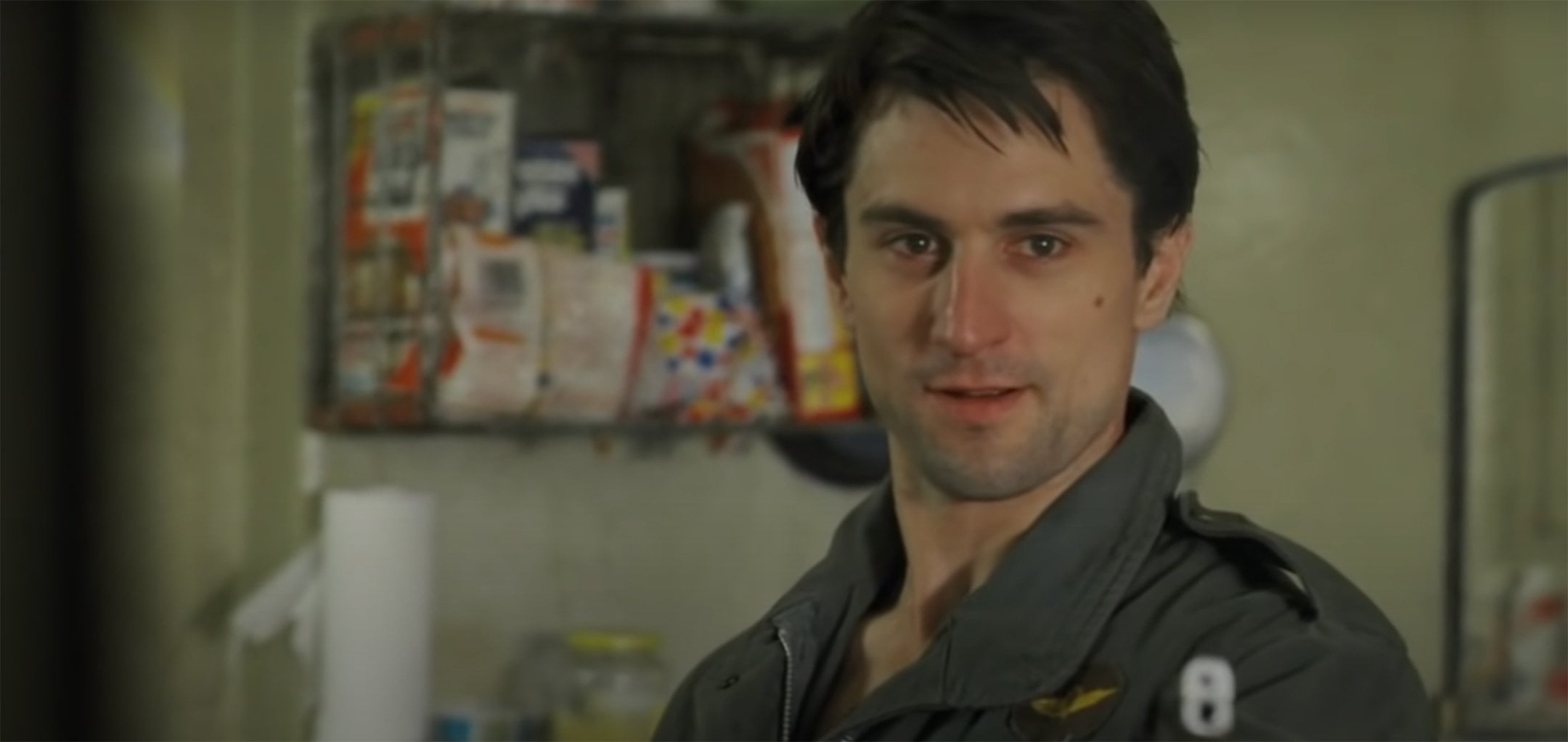 A man with dark hair wearing a green jacket stands indoors, looking slightly to the side and smiling. Shelves with food items and a paper towel roll are visible in the background.