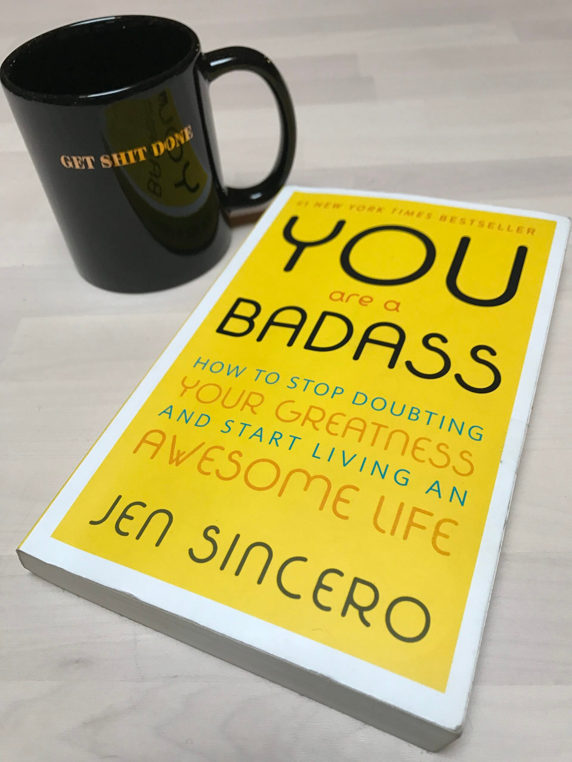 A black mug with gold text "GET SHIT DONE" sits next to the yellow cover of the book "You Are a Badass" by Jen Sincero on a light-colored surface.