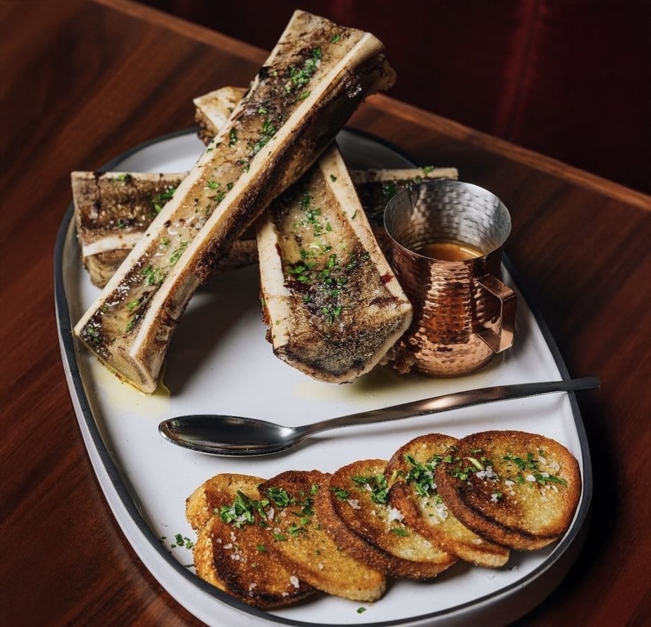 A plate with roasted bone marrow topped with herbs, alongside toasted bread slices, a small copper cup of sauce, and a spoon, set on a wooden table.
