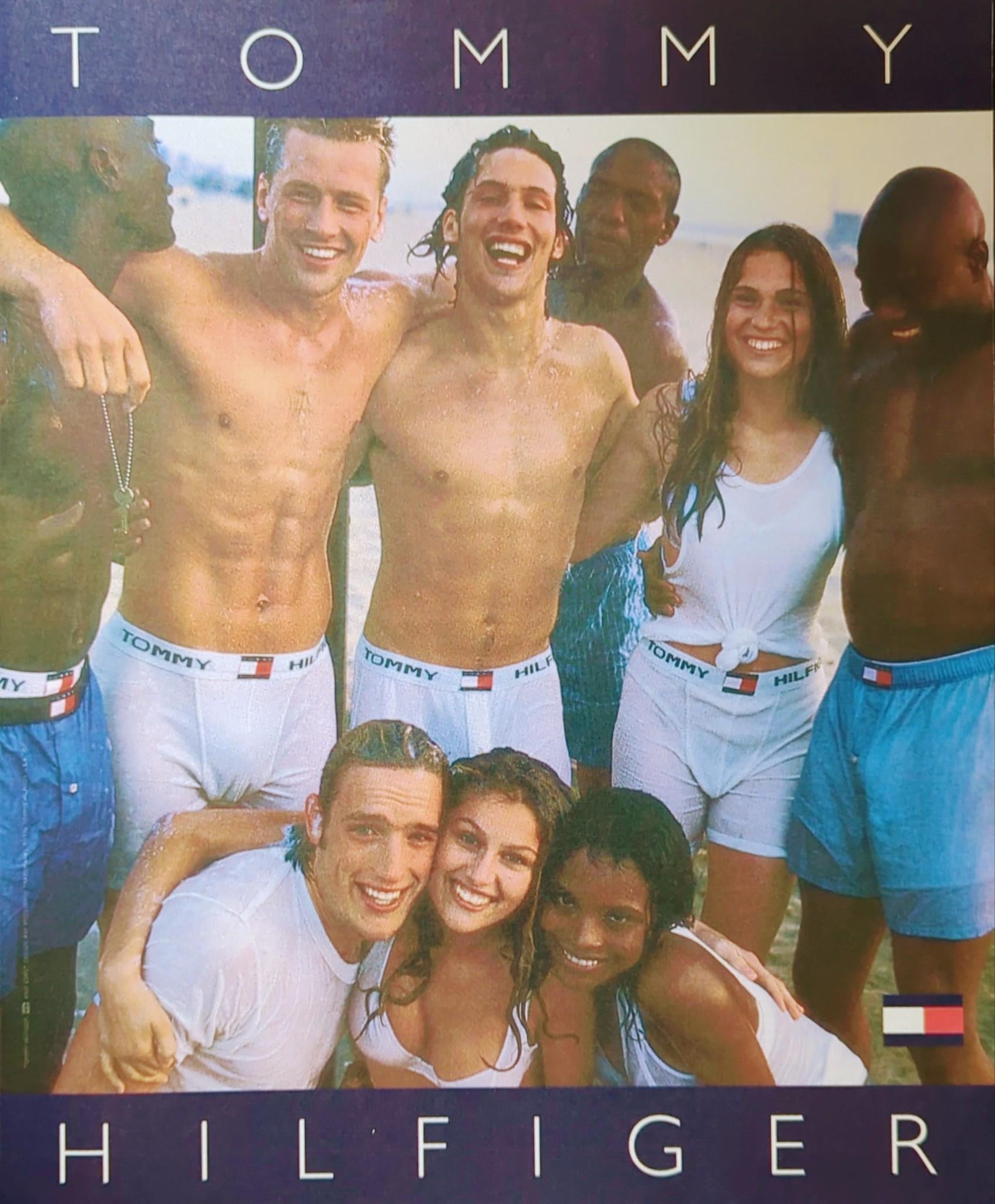 A group of smiling young adults in white Tommy Hilfiger underwear and tank tops pose together on a beach. They appear happy and carefree, with the Tommy Hilfiger logo visible above and below the image.