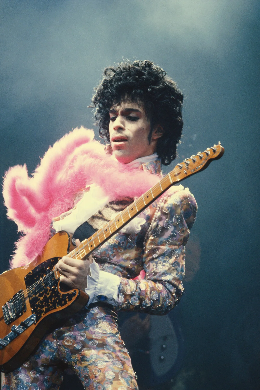 A musician in a patterned suit and pink feather boa plays an electric guitar passionately on stage under dramatic lighting.