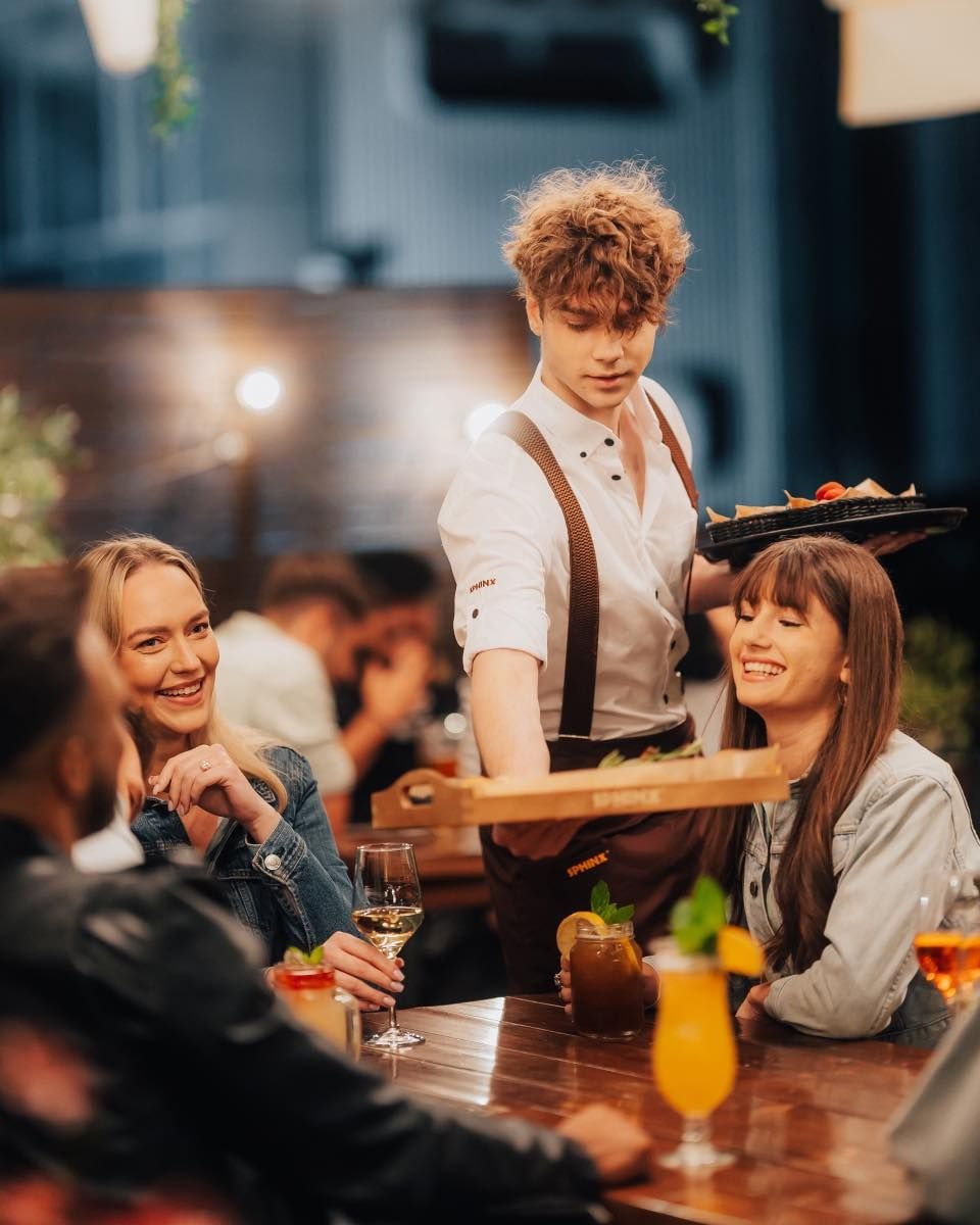A waiter serves food to a group of smiling friends sitting at a wooden table, enjoying drinks in a warmly lit, lively restaurant setting.