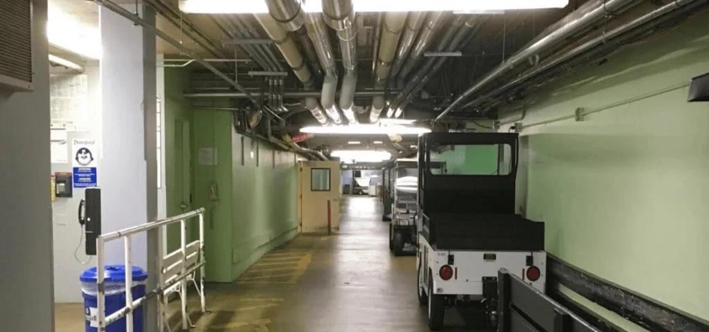 A basement corridor with exposed pipes on the ceiling, pale green walls, a small white utility vehicle parked by the side, safety signs, and a blue trash bin near a white railing.