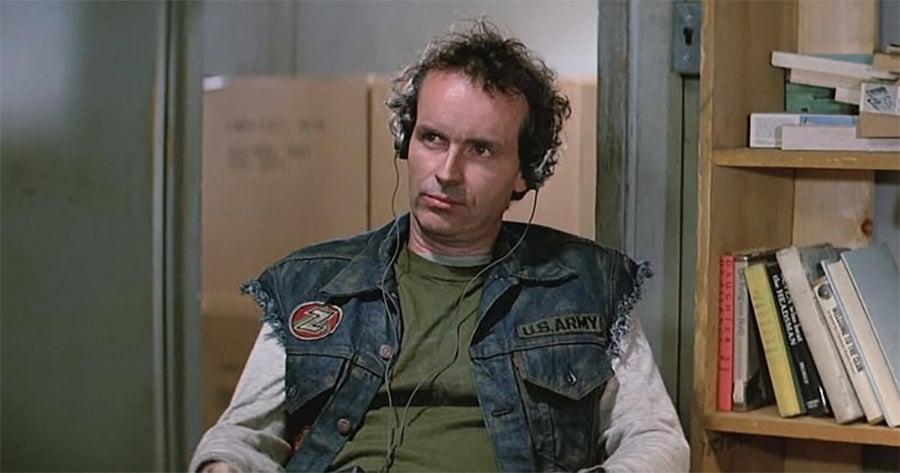 A man with curly hair wearing headphones, a green shirt, and a sleeveless denim vest with U.S. Army and Z patches sits indoors near shelves filled with books and papers.