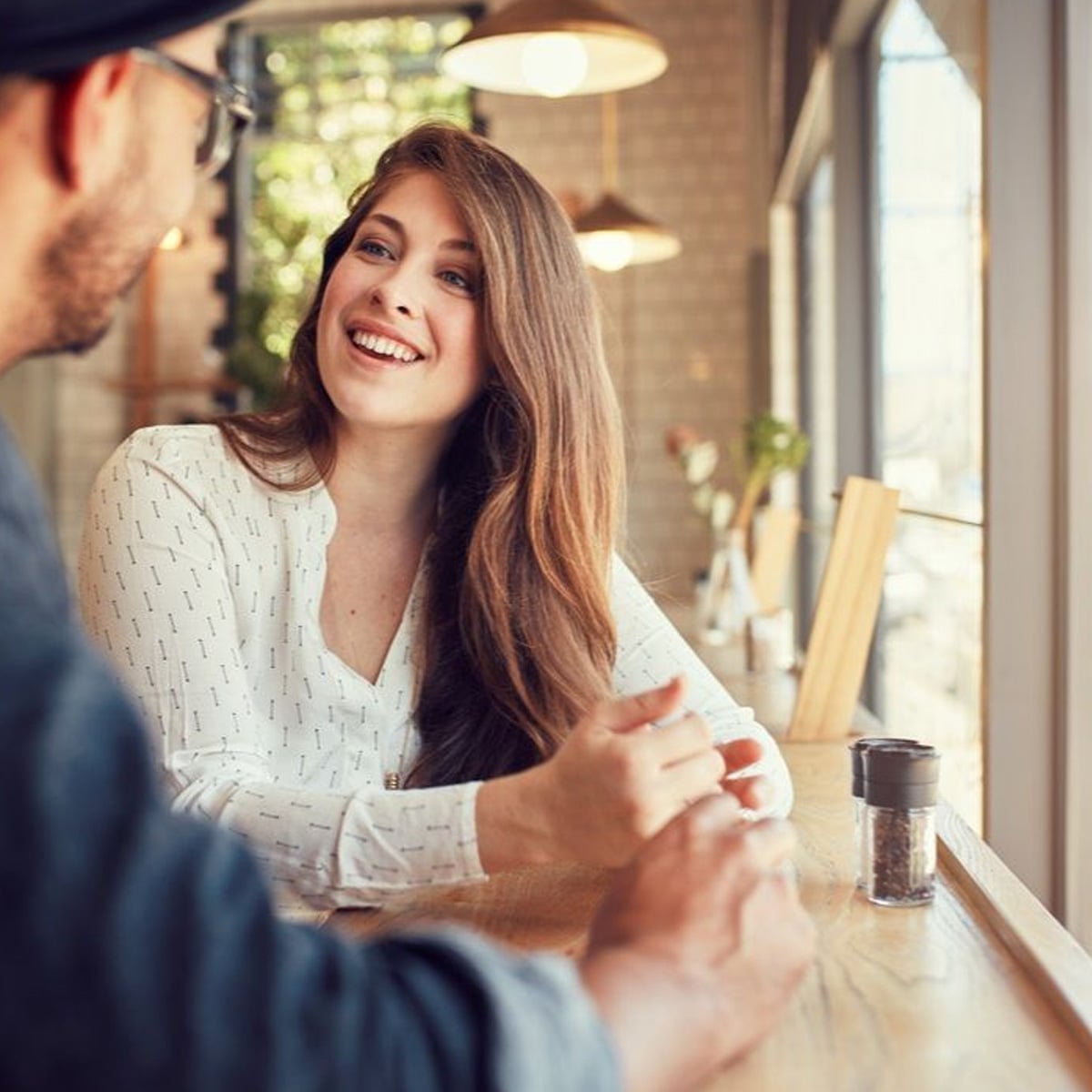 A woman with long brown hair smiles while sitting at a wooden café table, engaged in conversation with a man. Bright natural light streams through large windows in the cozy, modern setting.