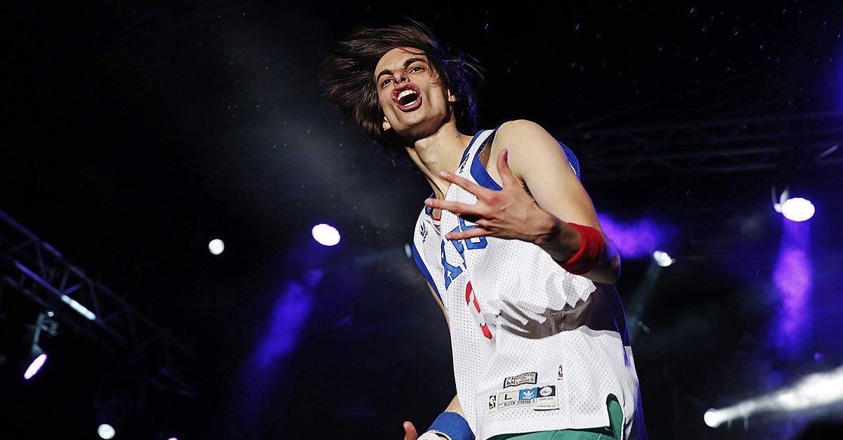 A young man with long hair and a white sports jersey makes a dynamic gesture onstage, with dramatic lighting and colorful spotlights in the background.