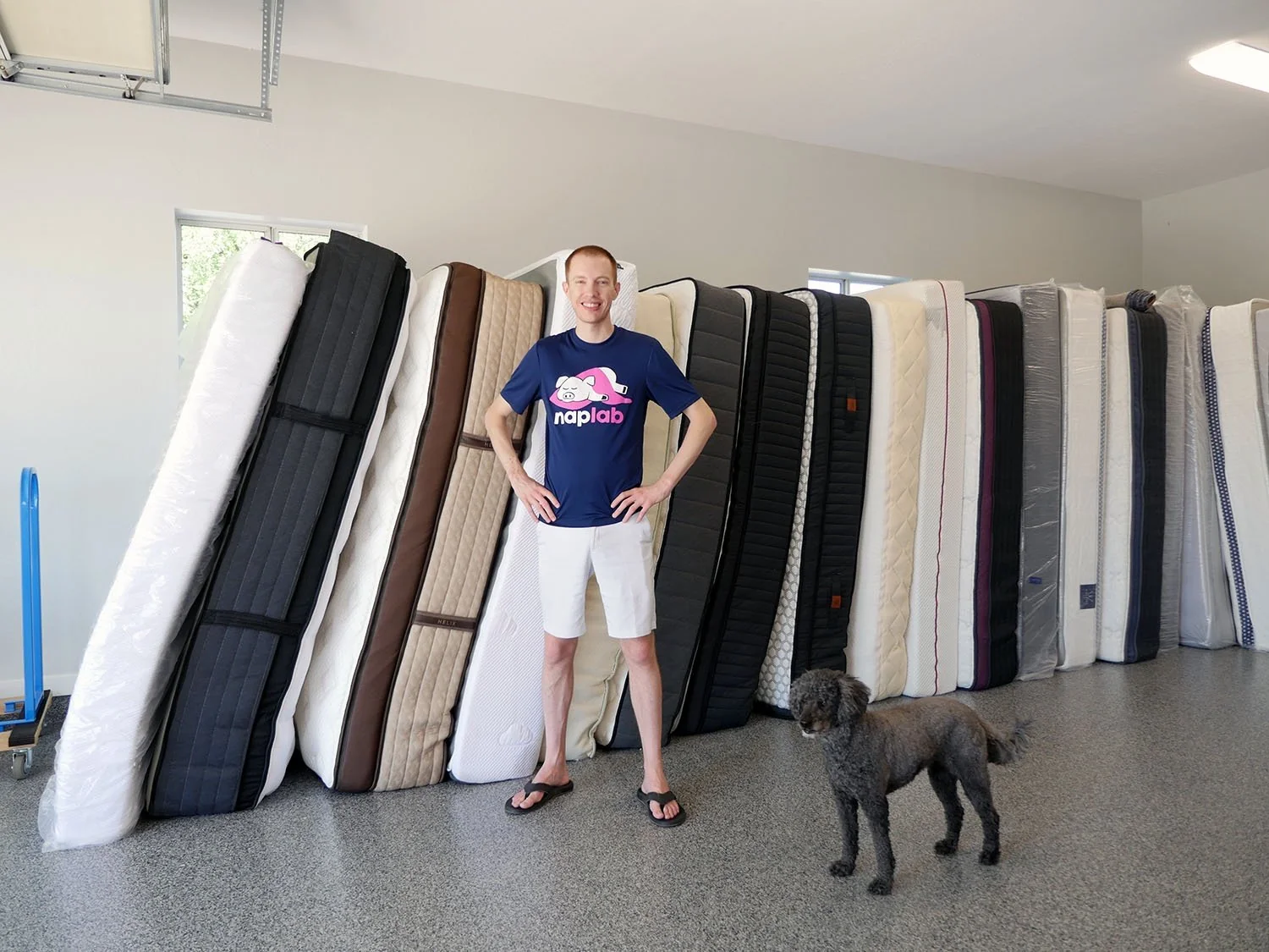A person stands in a garage next to a row of upright mattresses leaning against the wall, with a dog standing nearby on the gray floor. The person wears a blue shirt, light shorts, and flip-flops.