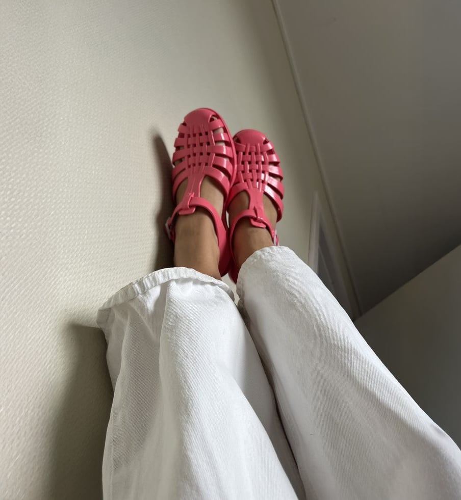 Person wearing loose white pants and bright pink jelly sandals, lying on their back with legs leaning against a textured off-white wall. The image is shot from a low angle, emphasizing the sandals.
