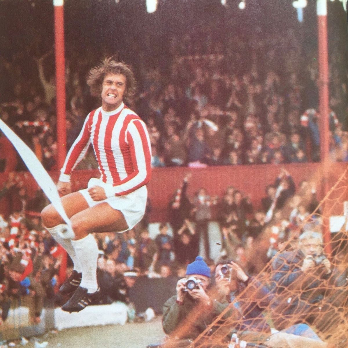 A soccer player in a red and white striped jersey and white shorts celebrates energetically after scoring, jumping with a triumphant expression as fans and photographers look on from the stands.