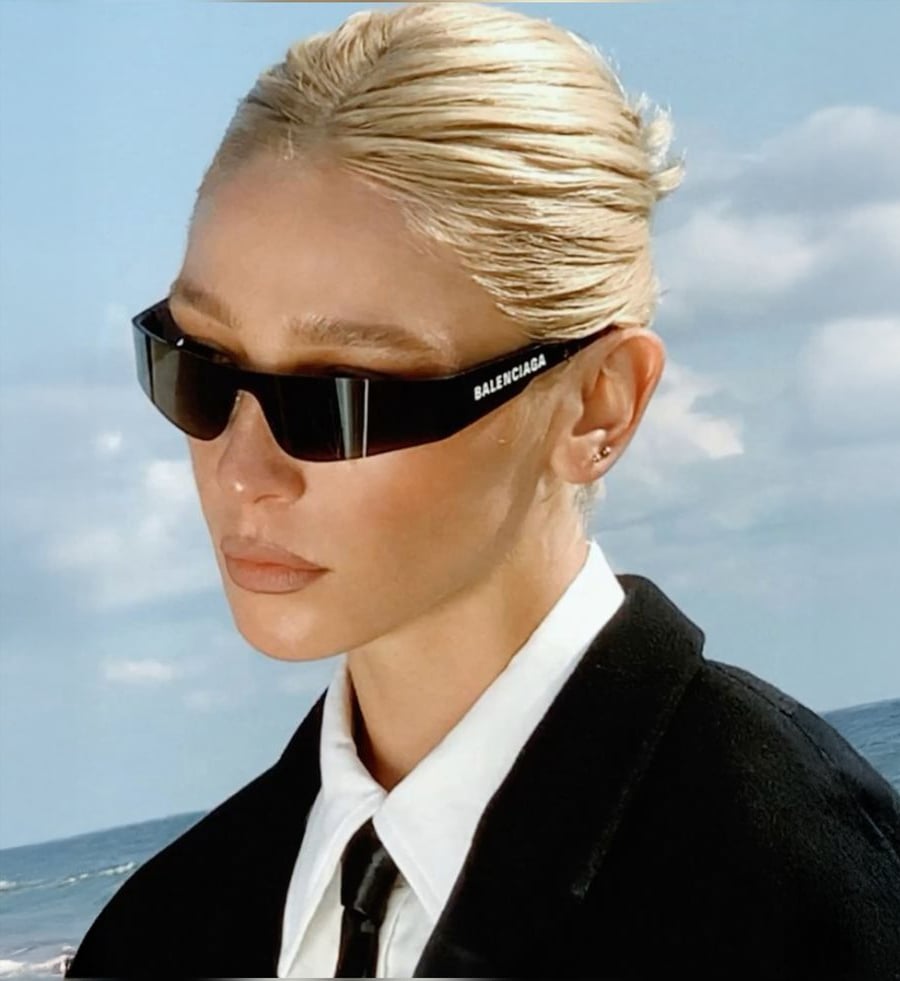 A person with slicked-back blonde hair wears black Balenciaga sunglasses, a white shirt, black tie, and black blazer, standing outdoors with blue sky and clouds in the background.