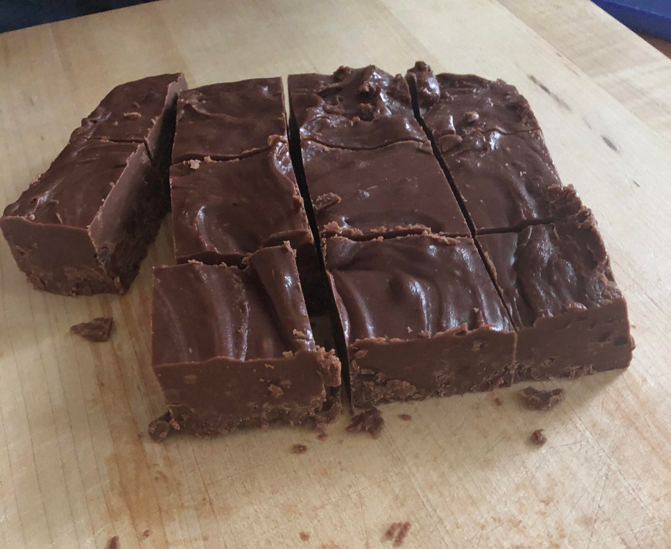 A batch of chocolate fudge cut into rectangular pieces sits on a wooden cutting board. The fudge has a smooth, glossy surface and slightly uneven edges.