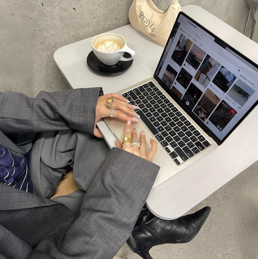 A person wearing a gray suit, rings, and black boots is sitting at a table using a laptop. A cup of coffee and a beige bag are next to the laptop. The screen shows a grid of photos.
