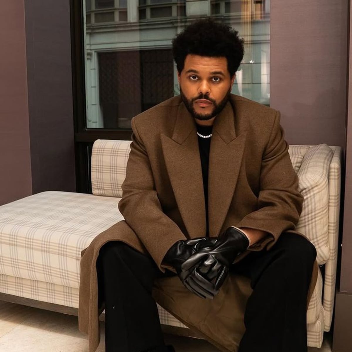 A man with short curly hair and a beard sits on a plaid couch, wearing a brown overcoat, black gloves, black pants, and a pearl necklace, looking directly at the camera in a modern indoor setting.