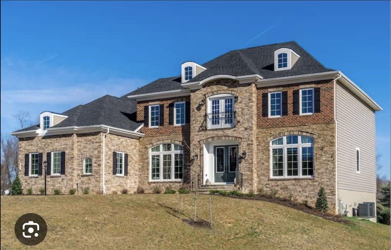A large two-story house with a combination of brick and siding exterior, tall windows, black shutters, a small balcony above the front entrance, and a sloped grassy lawn in front, under a clear blue sky.