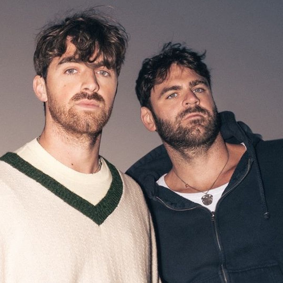 Two men with short brown hair and beards stand close together, looking at the camera. One wears a cream sweater with green trim, and the other wears a dark hoodie over a T-shirt. The background is plain and dimly lit.