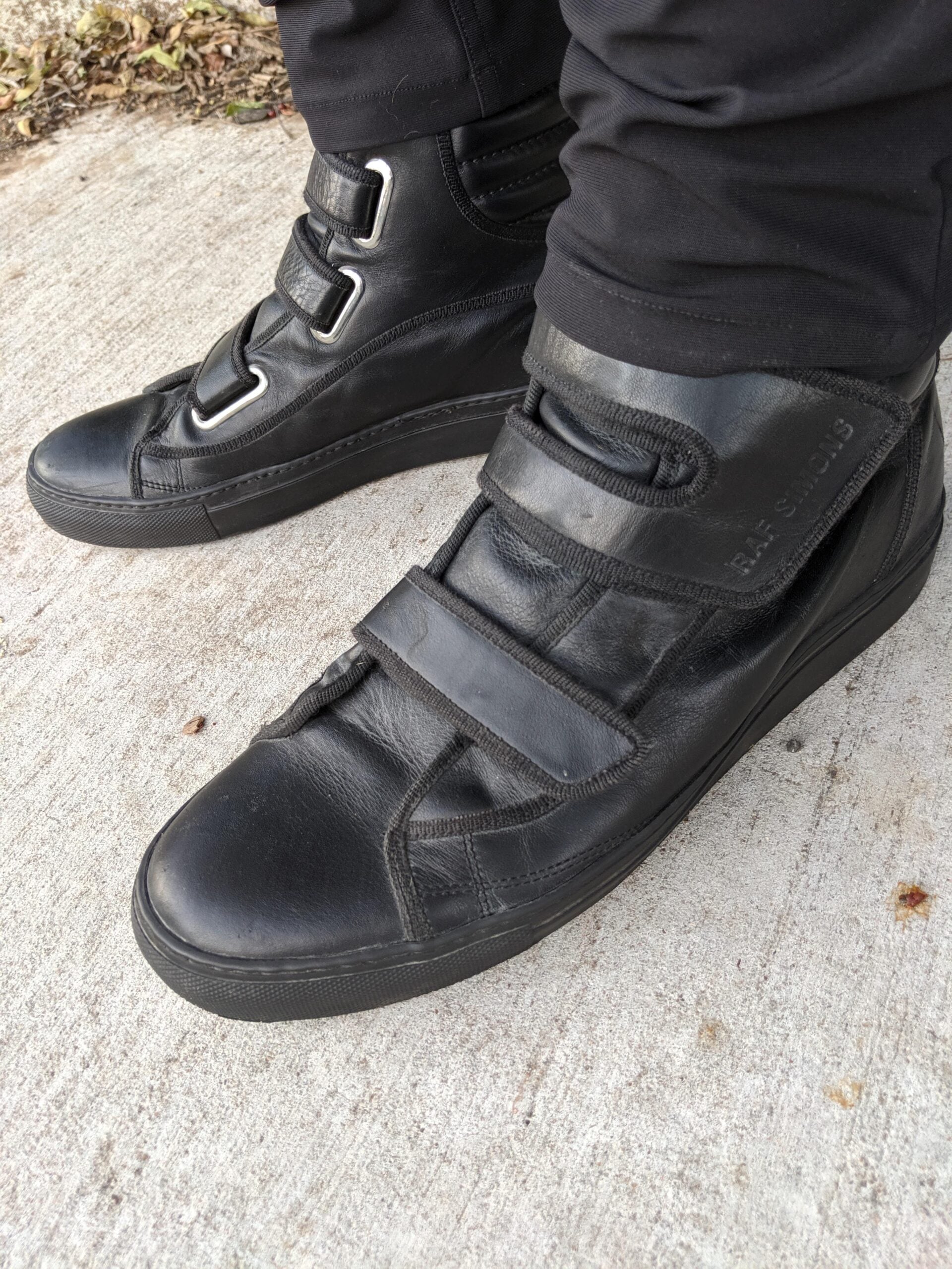 A person wearing black high-top sneakers with Velcro straps, standing on a concrete sidewalk. The shoes have a sleek design and are paired with black pants.