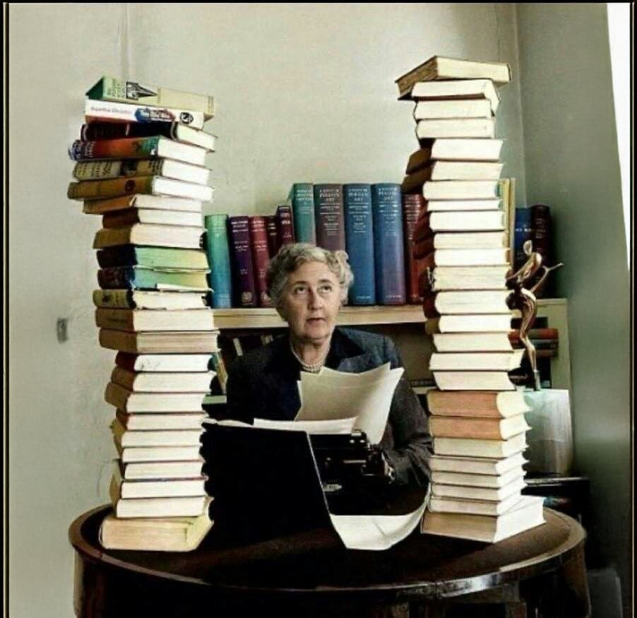 An older woman sits at a desk with a typewriter, surrounded by two tall, leaning stacks of books. Shelves filled with colorful books are in the background. She appears focused on her work.