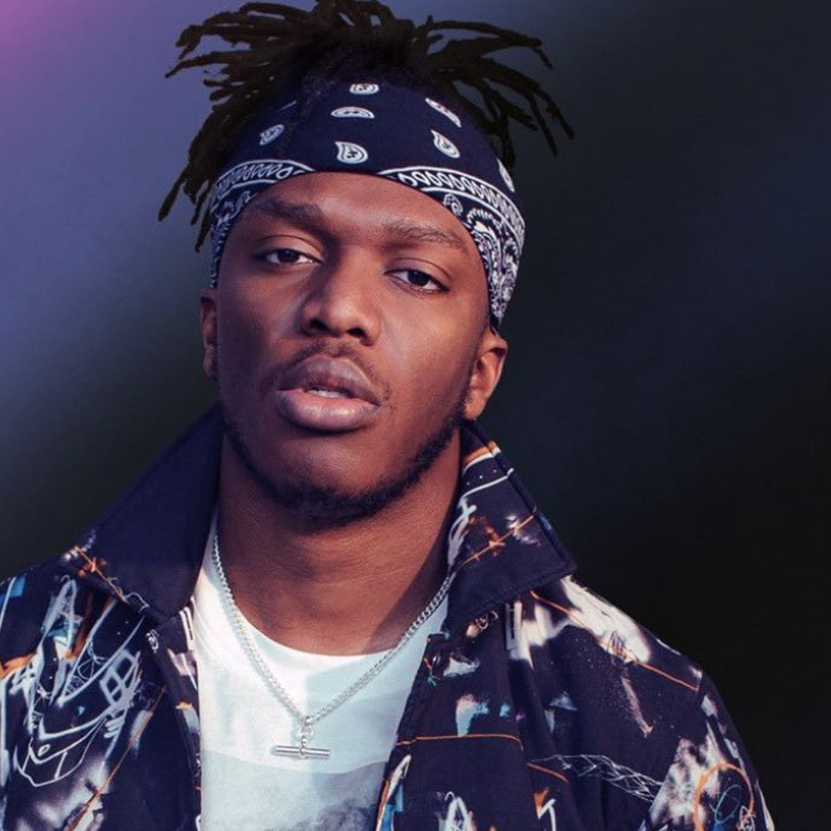 A man wearing a patterned bandana and jacket, layered over a white t-shirt, with a chain necklace, looking ahead with a neutral expression against a dark, blurred background.