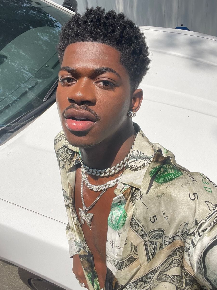 A young man with short curly hair takes a selfie outdoors by a white car. He wears a patterned shirt with dollar bill prints, layered silver chains, and a butterfly pendant, with sunlight highlighting his face.