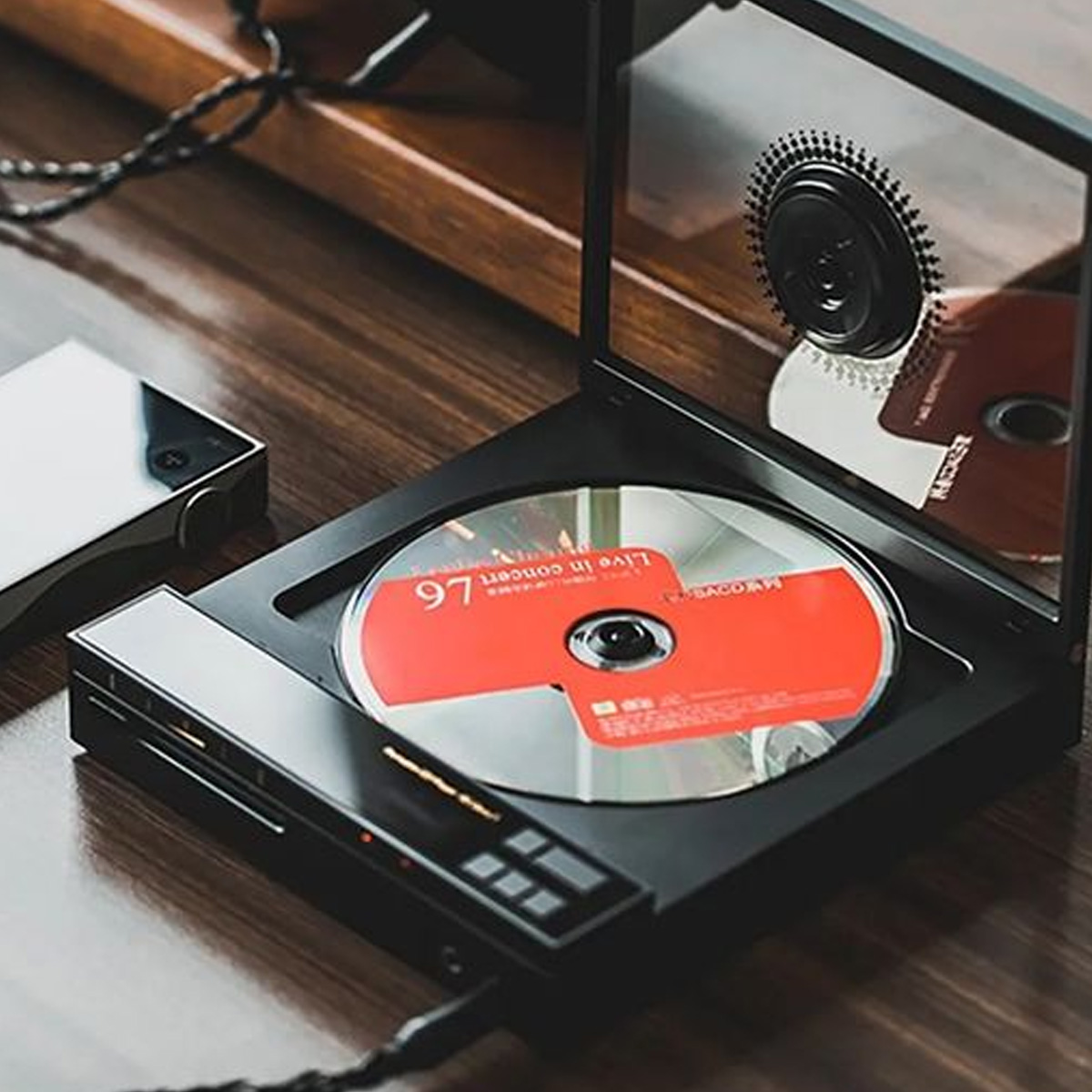 A red CD is placed inside a sleek, open black CD player on a wooden surface. The CD player has a transparent lid with a visible spindle and some control buttons on its front panel.