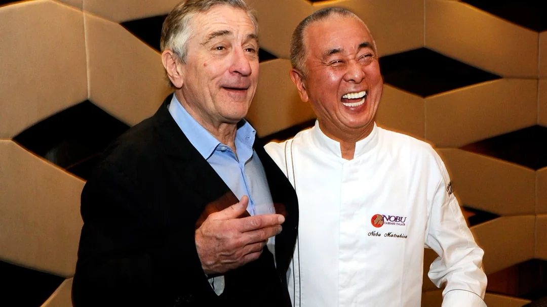 Two men stand together smiling; one wears a dark suit and light blue shirt, while the other, in a white chef’s coat with “Nobu” and “Nobu Matsuhisa” embroidered, holds his arm around the suited man in front of a patterned wall.