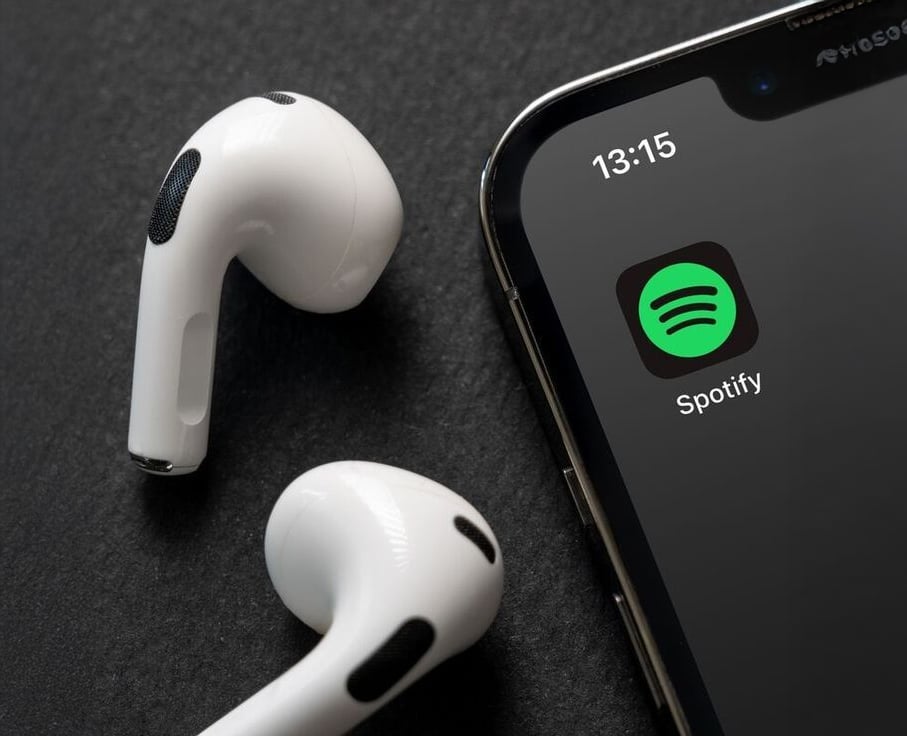 Close-up of wireless white earbuds next to a smartphone displaying the Spotify app icon on its screen. The phone shows the time as 13:15 against a dark background.