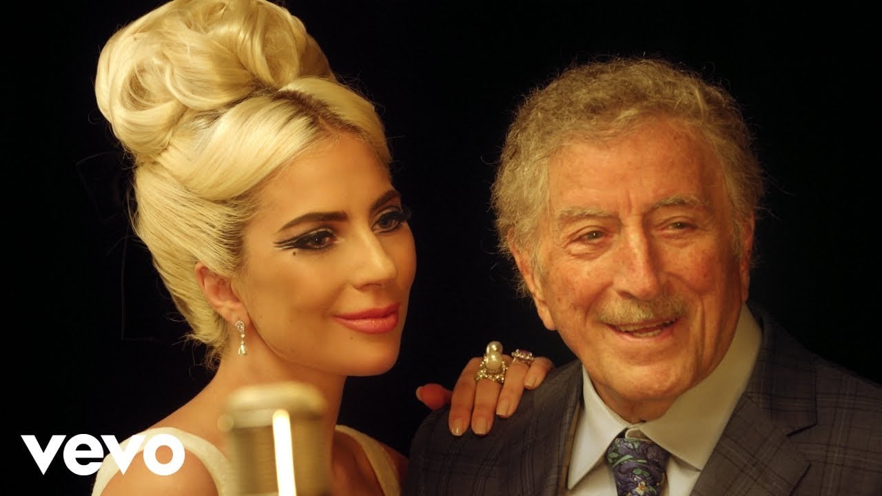 A woman with blonde hair in an elegant updo and a man with gray hair and a mustache smile together in front of a microphone, dressed in formal attire against a dark background.