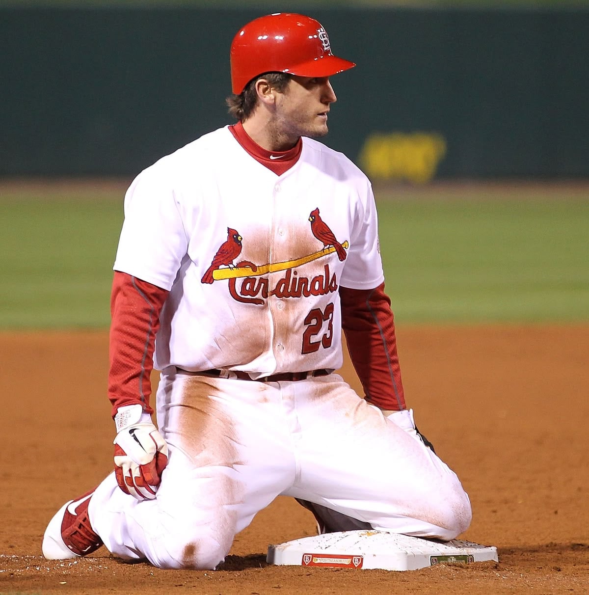A St. Louis Cardinals baseball player, wearing a white uniform and red helmet, kneels on the base path with one knee on second base during a game. The field is dirt and grass, and the player's uniform is dirty.
