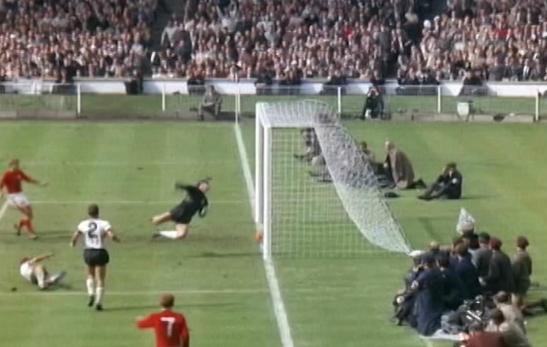 A dramatic moment in a soccer match as a player in white shoots at goal; the goalkeeper dives to make a save, while players and the crowd watch intently behind the net in a packed stadium.