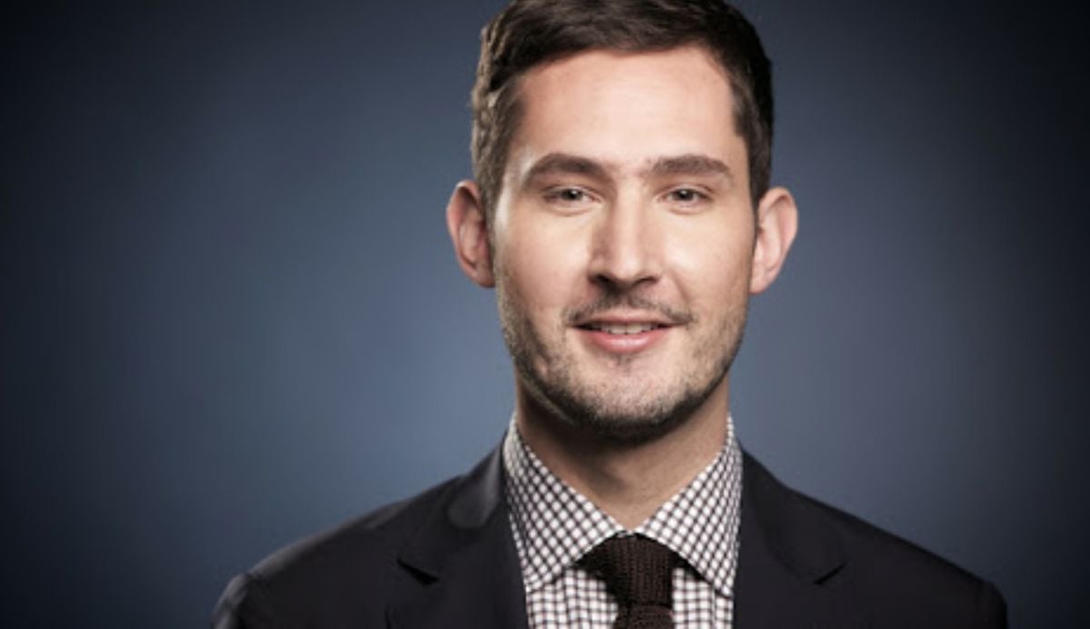 A man with short dark hair and light stubble is smiling slightly. He is wearing a dark suit, a checkered shirt, and a dark tie, posed against a plain dark background.
