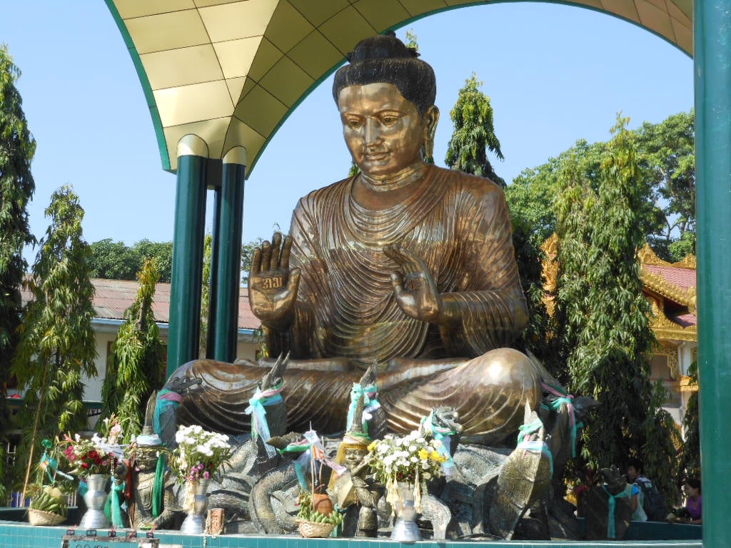 A large bronze Buddha statue sits under a pavilion, surrounded by offerings of flowers and small sculptures, with trees and buildings in the background on a sunny day.