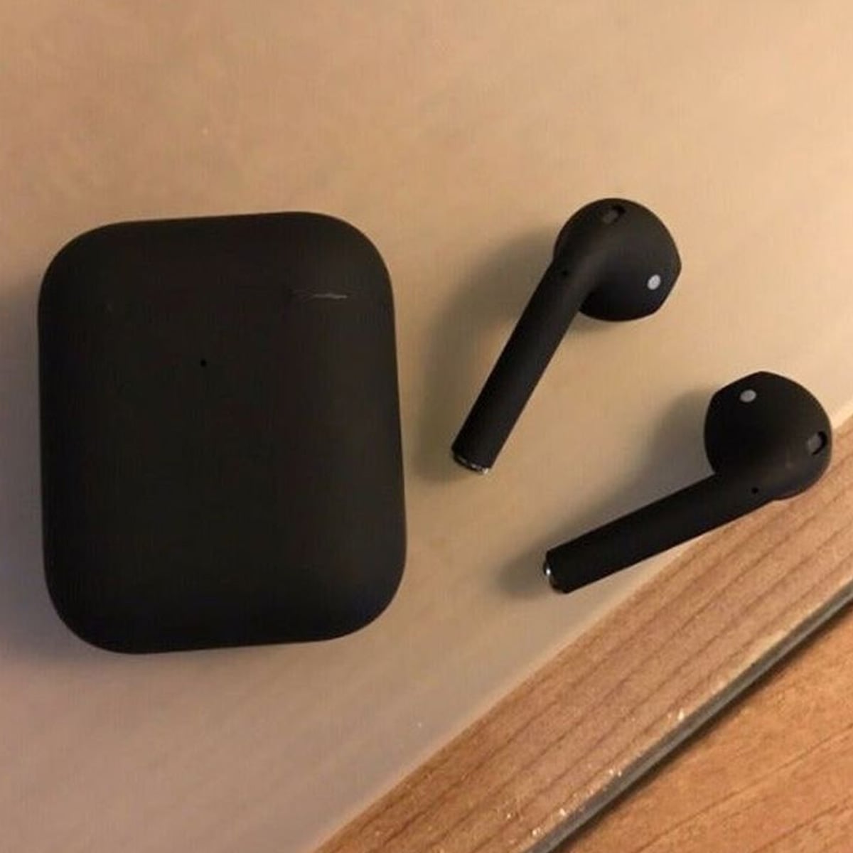 A pair of black wireless earbuds and their matching charging case rest on a light-colored surface next to a wooden edge.