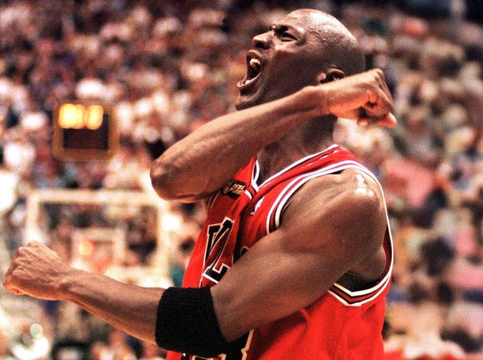 A basketball player in a red jersey celebrates passionately on the court, clenching his fists and yelling, with a blurred crowd in the background.