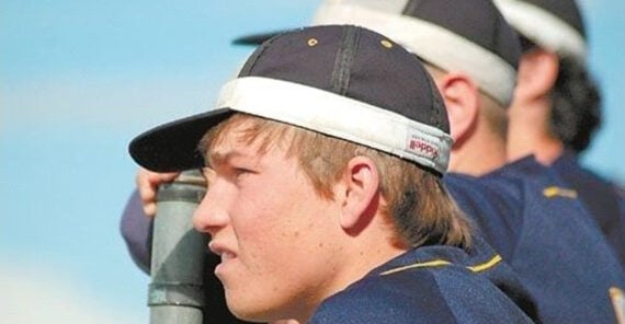 A young baseball player in a navy cap and jersey leans against a fence, looking out onto the field. Another player in similar attire is visible in the background.