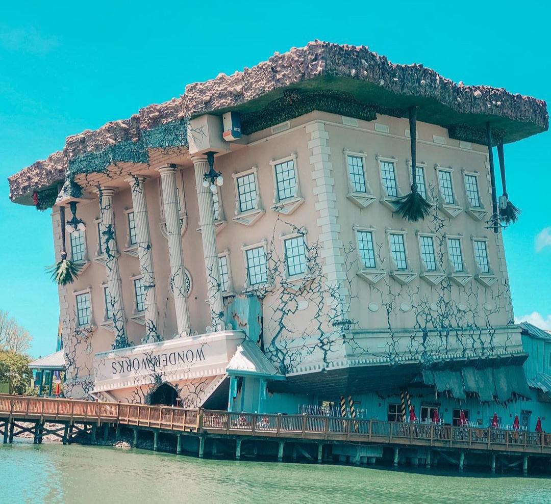 A large upside-down building with classical columns and windows, called WonderWorks, appears to have crash-landed, with its roof smashed into the ground, set beside a body of water on a sunny day.