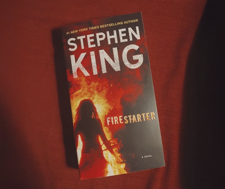 A book titled "Firestarter" by Stephen King rests on a red surface. The cover features a silhouette of a girl standing in front of fiery flames.