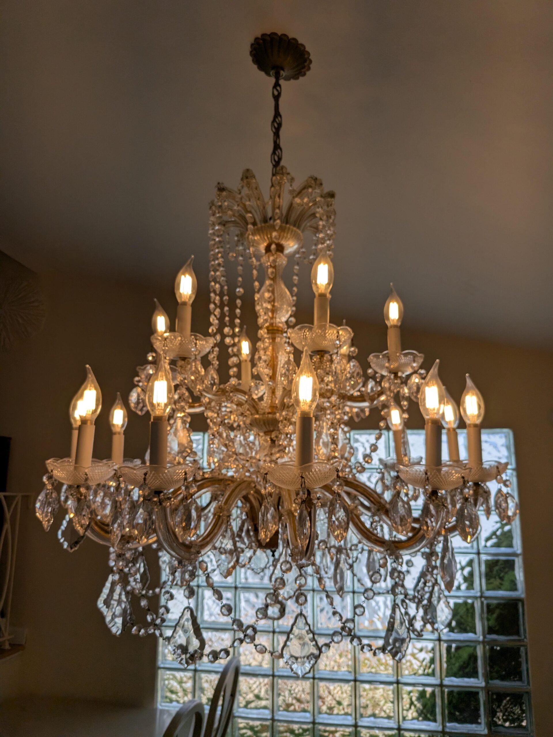 A crystal chandelier with lit candle-shaped bulbs hangs from the ceiling. Glass prisms and beads decorate the fixture. In the background, large glass block windows let natural light into the room.