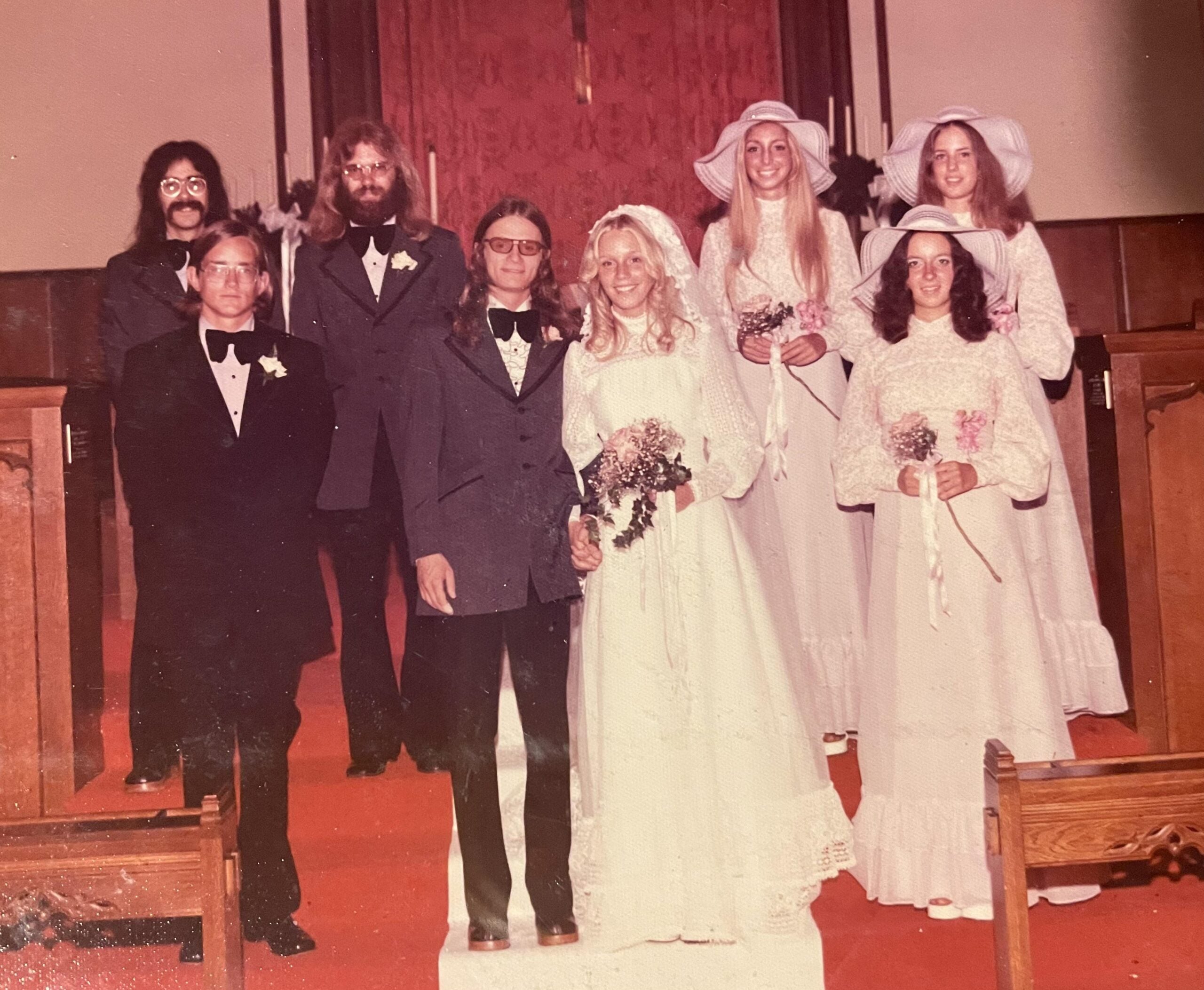 A vintage wedding party poses inside a church. The bride and groom stand at the center, surrounded by bridesmaids in white dresses and large hats, and groomsmen in suits, all on a red carpeted aisle.