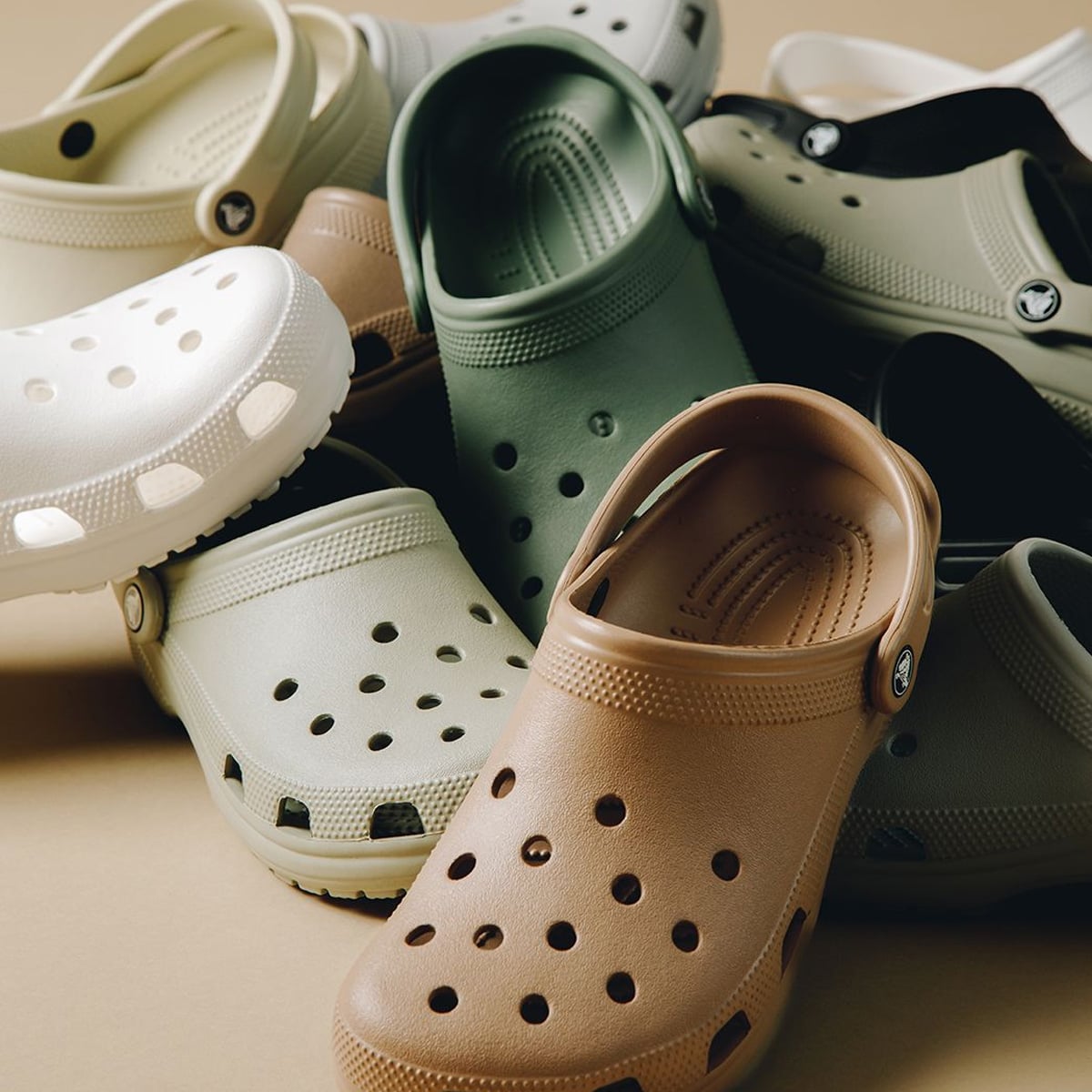 A pile of rubber clogs in neutral colors including green, brown, white, and beige, arranged on a beige surface. The clogs have ventilation holes and adjustable heel straps.