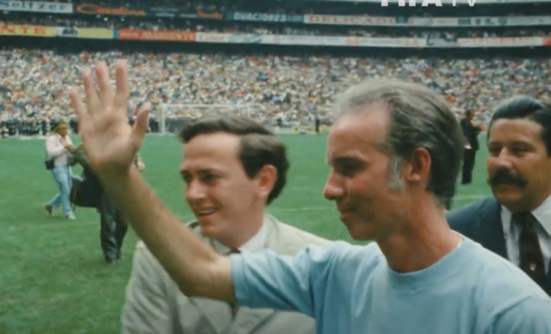 A man in a light blue shirt waves to a cheering crowd in a large stadium, with two other men standing beside him on the grass. The stands are filled with spectators.